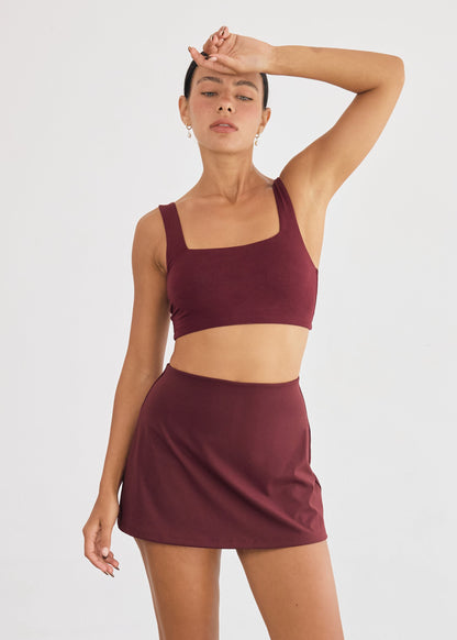 Woman wearing a maroon sports bra and skirt set on a plain background