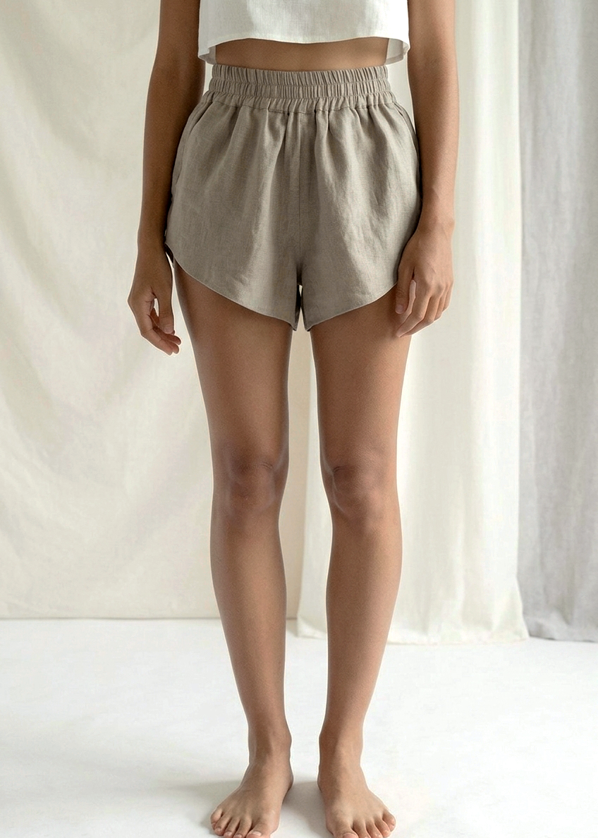 A model from the waist down wearing light beige linen shorts with an elastic waistband, standing barefoot against a draped white fabric background.