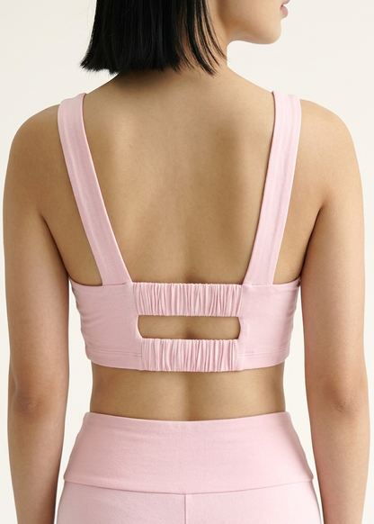 A woman shown from the back wearing a light pink sports bra with wide straps and a horizontal cutout at the center, paired with matching high-waisted leggings against a plain light background.