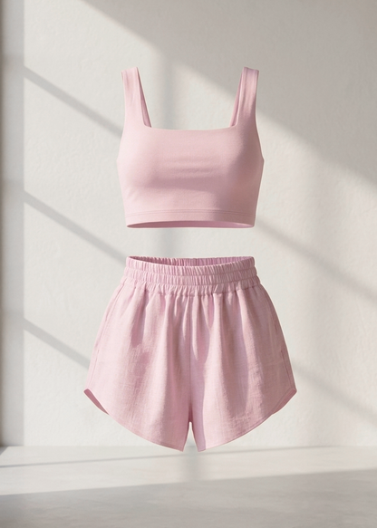 A two-piece lounge set in light pink, featuring a sleeveless scoop-neck crop top and matching high-waisted shorts with an elastic waistband, shown floating against a sunlit white wall.